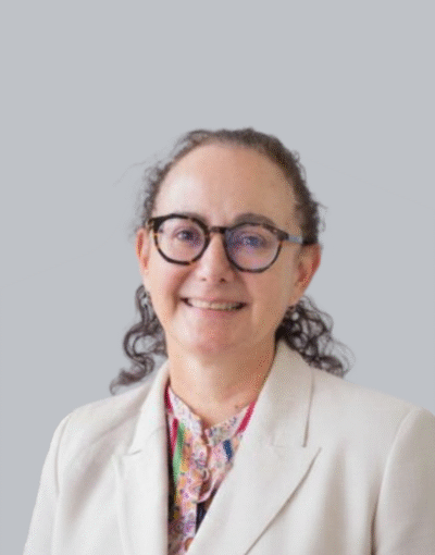 A woman wearing glasses and a beige suit, smiling, against a grey background.