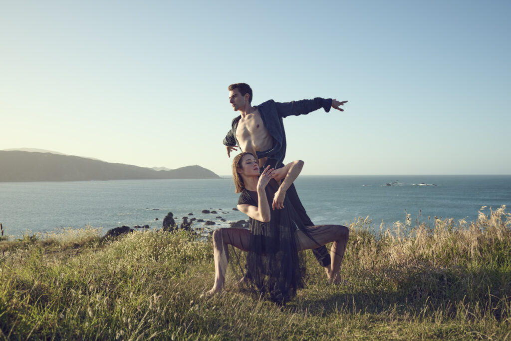 Two contemporary dancers pose dramatically on grassy cliffs by the ocean at sunset, with distant hills under a clear sky. The male dancer stands with one arm outstretched, while the female crouches gracefully in front of him.