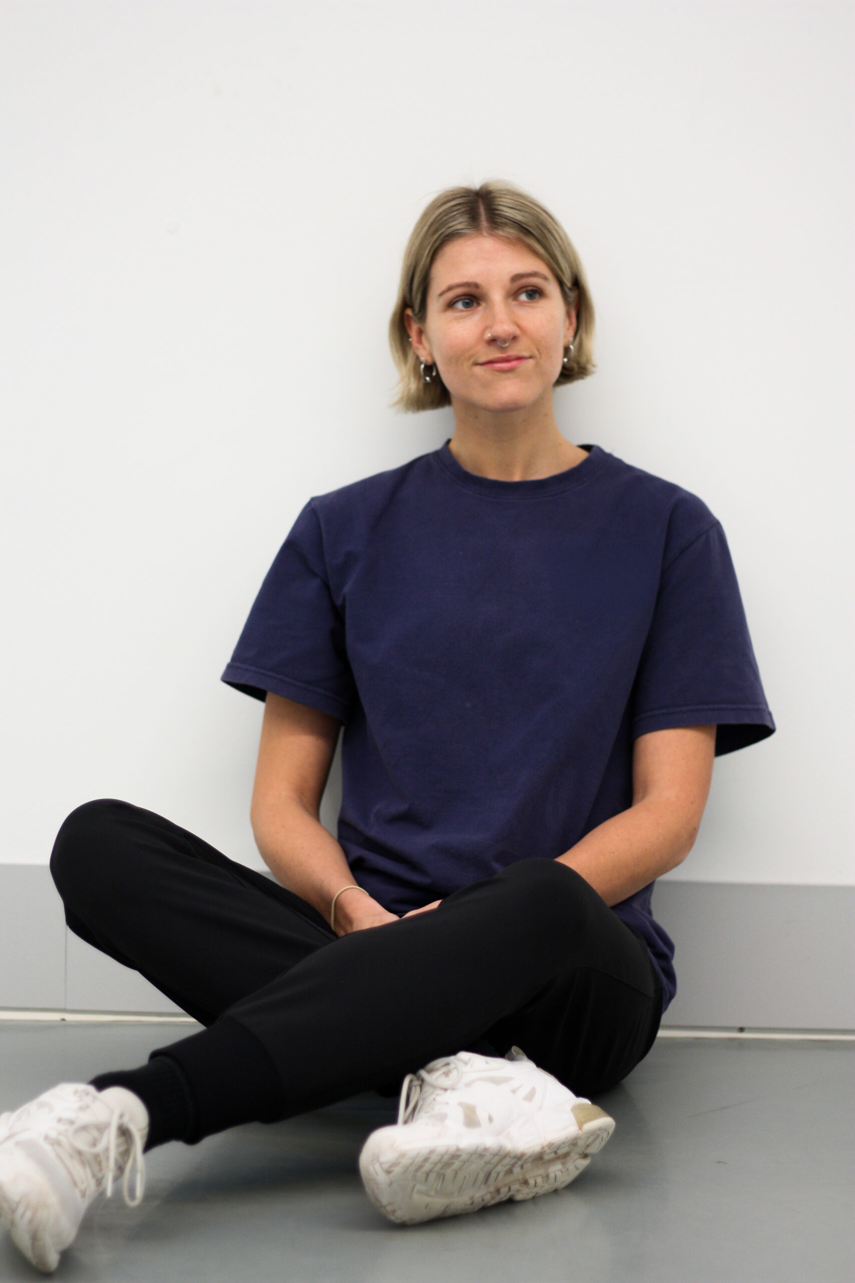 Lauren has blonde short chin length hair and sits against a white wall with a neutral expression. She wears a blue shirt, black trousers and white trainers.