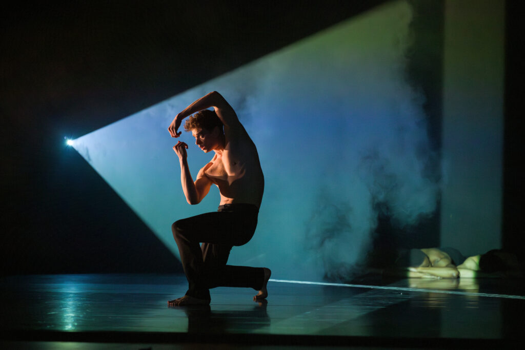 Josh, a masculine dancer with no shirt and black pants crouches as a blue and greem prism breaks through the darkness and frames him.