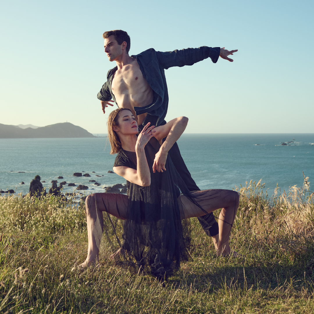 Two dancers on a grassy knoll, wind blowing, with the sea in the background.