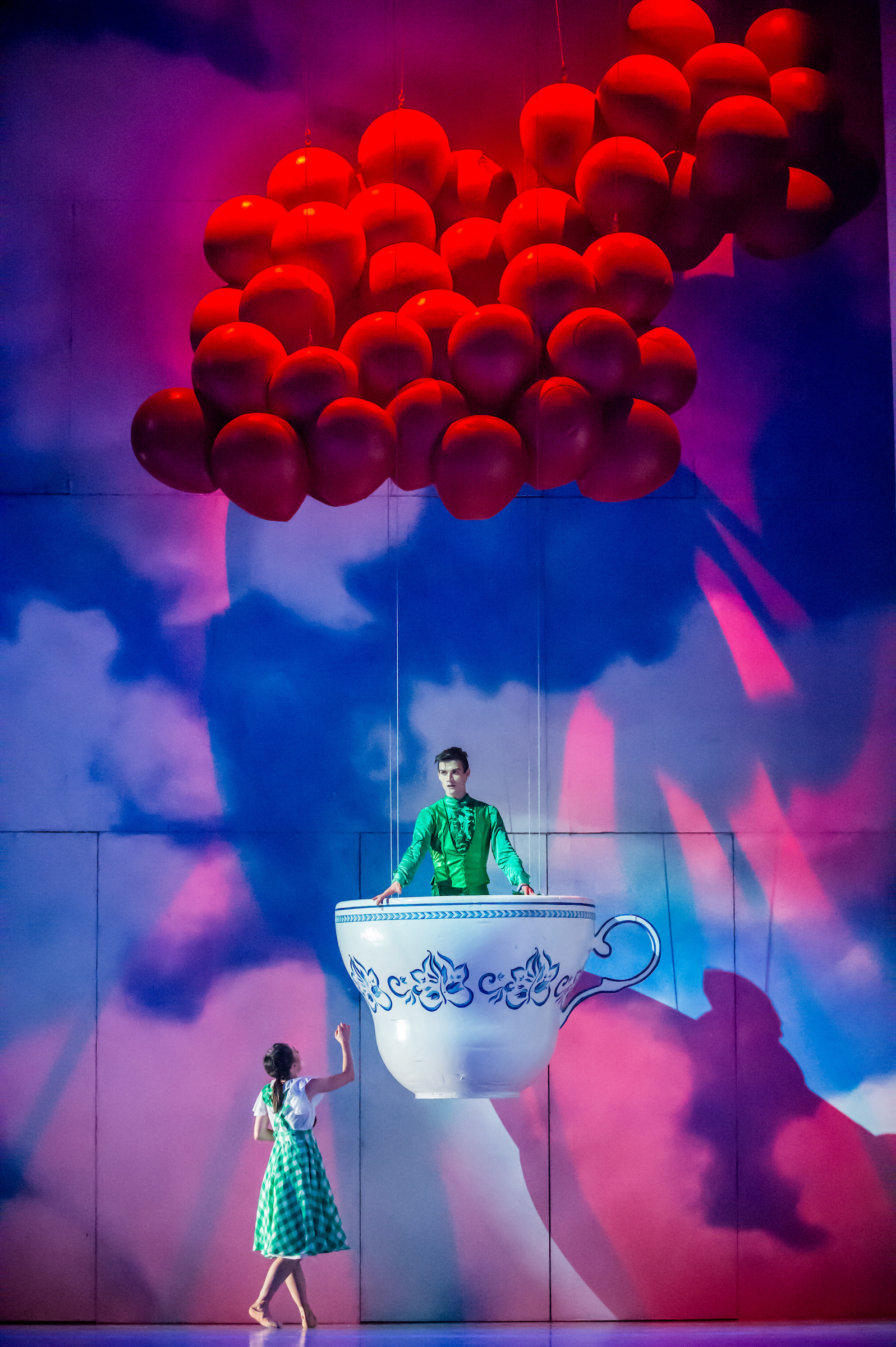 A woman in a green checkered dress stands beside a giant teacup containing a man in a green shirt, with a large cluster of red balloons hanging above them against a colorful, whimsical backdrop.