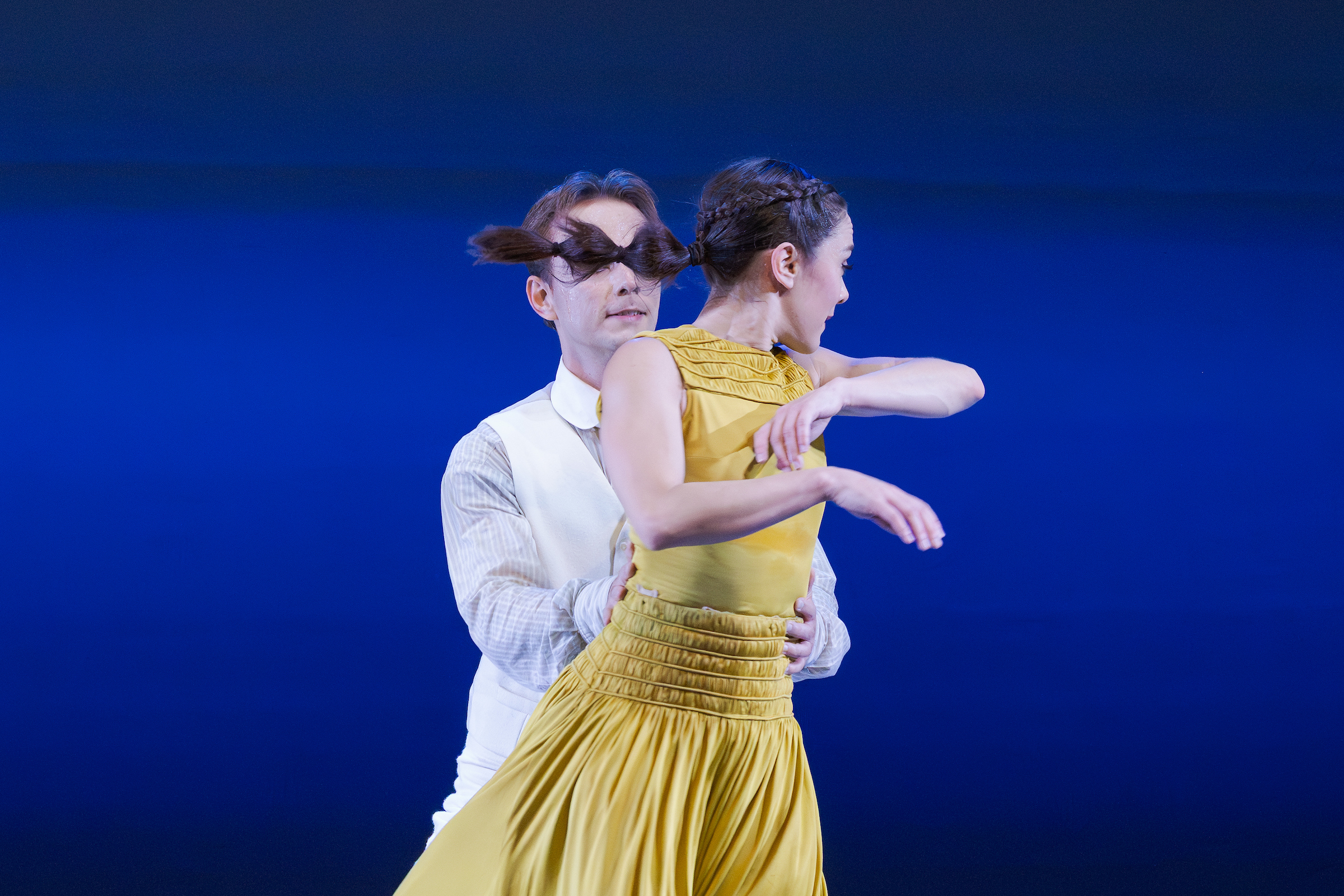 A man and woman dance together against a blue background. The woman wears a yellow dress and is spinning, her hair flying. The man, in a white shirt and vest, supports her from behind, both focused on their movement.
