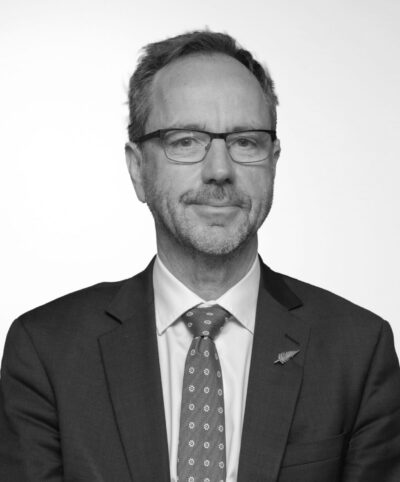 A black and white photo of John Ryan, a pākeha man wearing glasses, a dark suit with a tie and white shirt.